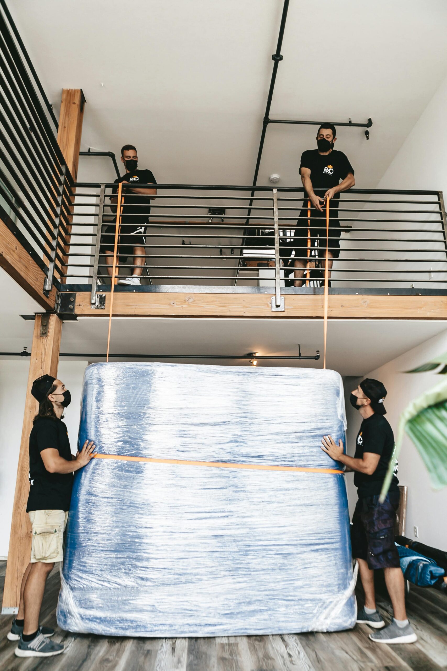 Four movers carefully lift a large wrapped object using straps indoors.