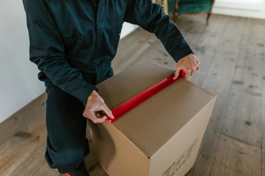 Male courier sealing a cardboard box with red tape, indoor wooden floor setting.