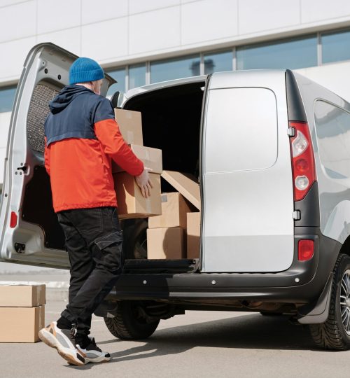 Man in colorful jacket loading cardboard boxes into a van outside an office building.