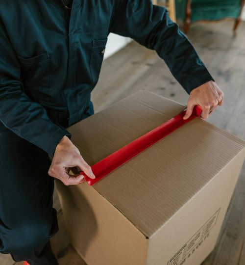 Male courier sealing a cardboard box with red tape, indoor wooden floor setting.